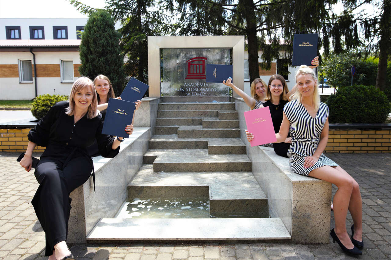 Studenci trzymają prace dyplomowe obok fontanny z logo Uczelni